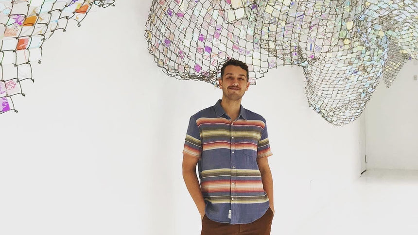 A man wearing a blue short-sleeve button-up shirt with horizontal multicolor stripes (red, orange, yellow, green) and brown pants, standing in a white room with a large net-like art installation hanging above him.